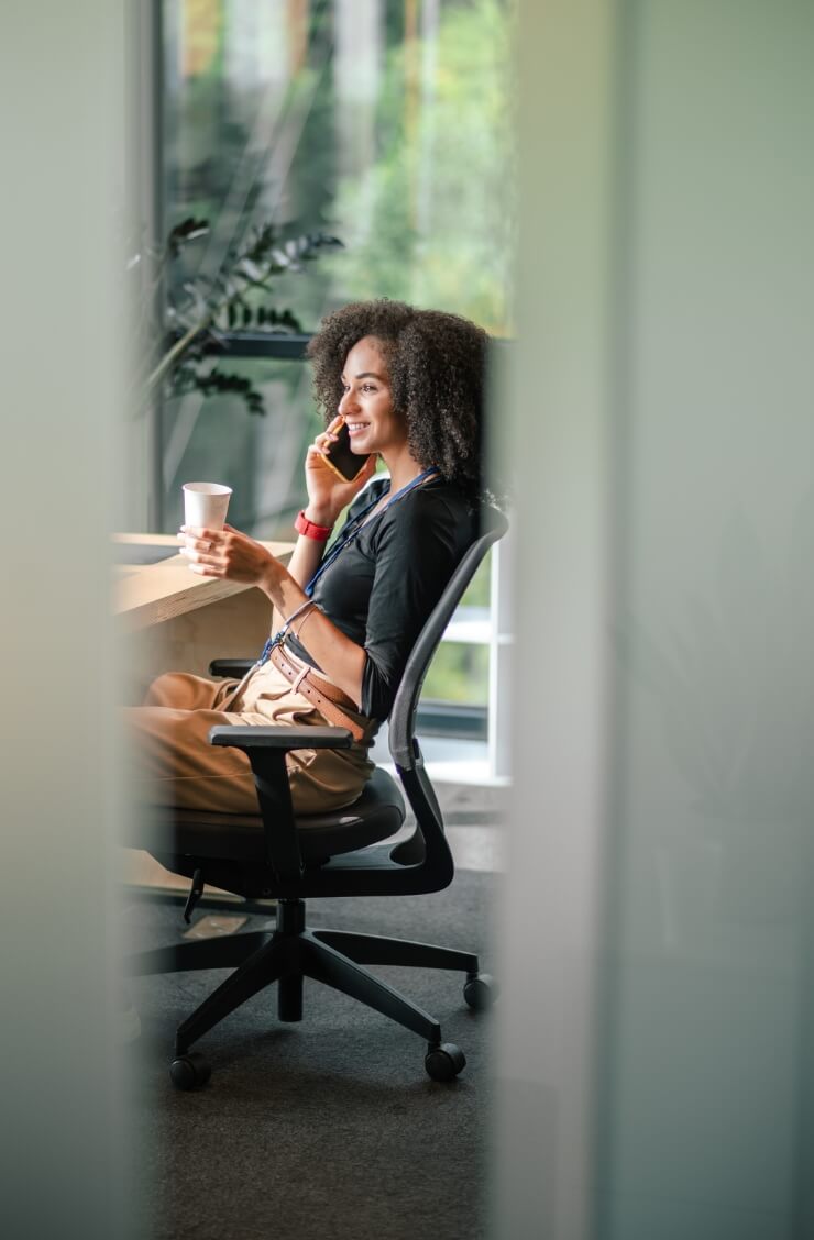 Mujer/Hombre sentado en una silla ergonómica, hablando por teléfono con cara sonriente mientras sujeta un vaso de cartón.